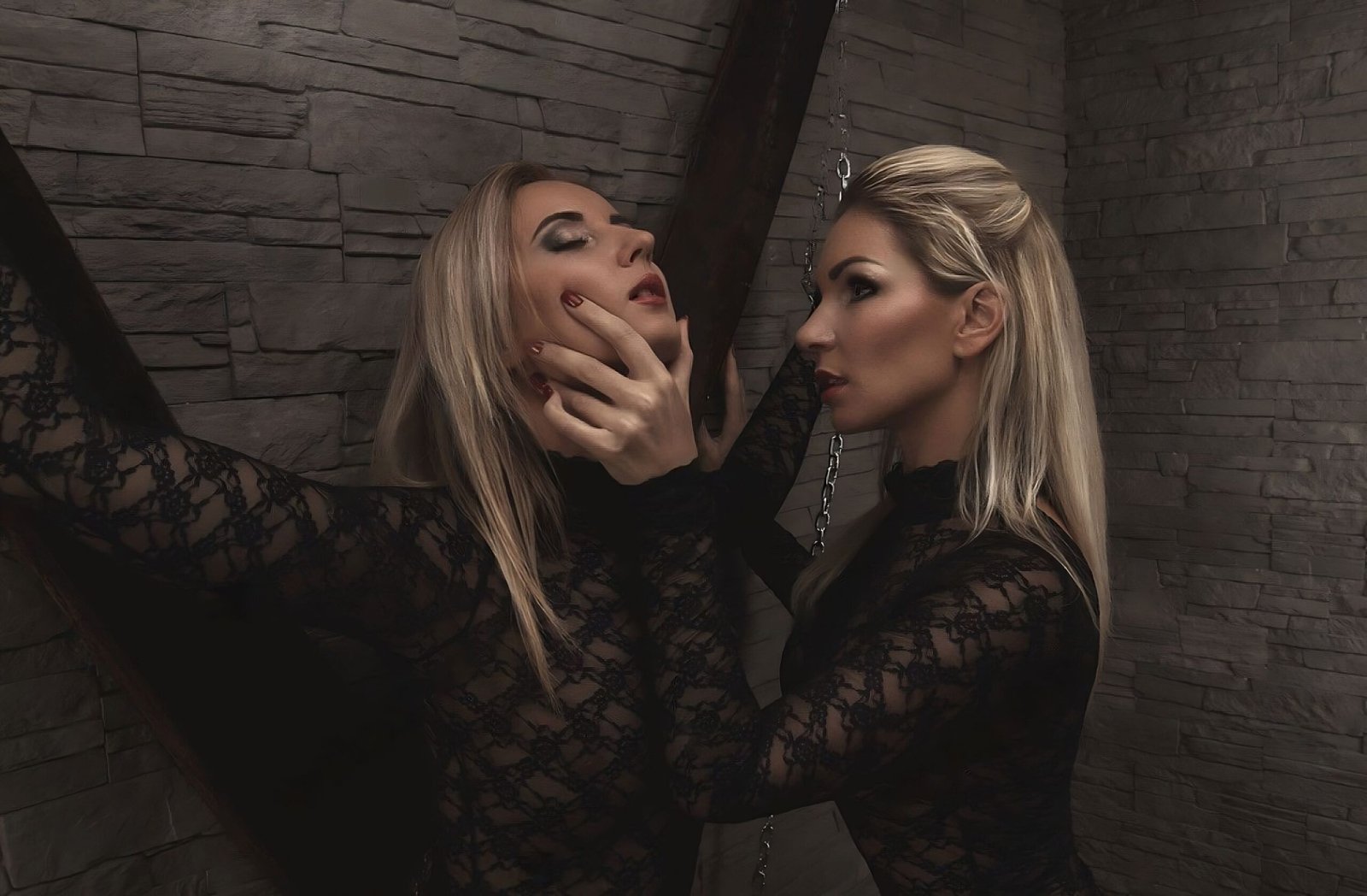 Two women with blonde hair, dressed in black lace, sharing an intimate moment against a stone wall.