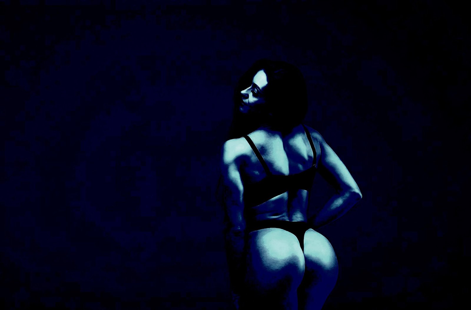 A woman standing in a dark room, wearing black lingerie, confidently looking back at the camera.