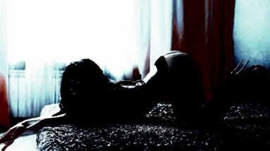 A woman in a seductive pose on a bed, lit by the soft glow of daylight coming through sheer curtains.