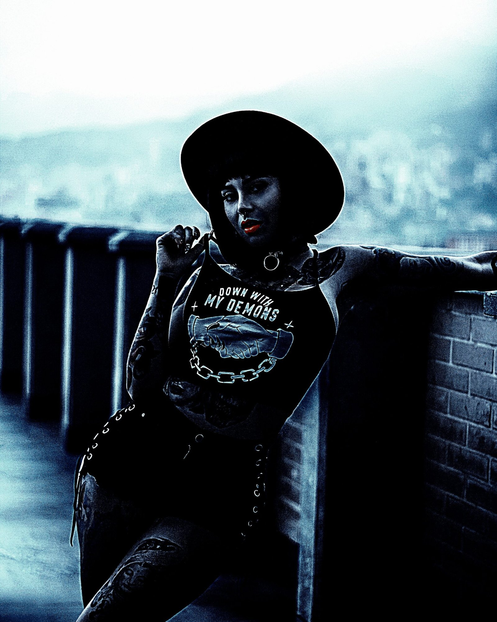 Gothic woman with tattoos and piercings, wearing a hat and "Down with My Demons" tank top, posing outdoors.