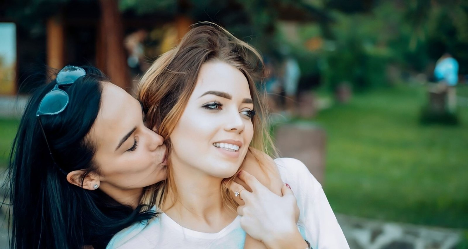 Two women, one with dark hair kissing the cheek of a blonde woman, sharing an intimate and happy moment outdoors.