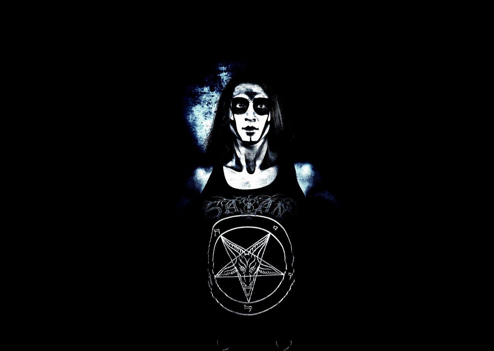 A person wearing a black tank top with a prominent pentagram symbol, face painted in black and white, standing against a dark background.