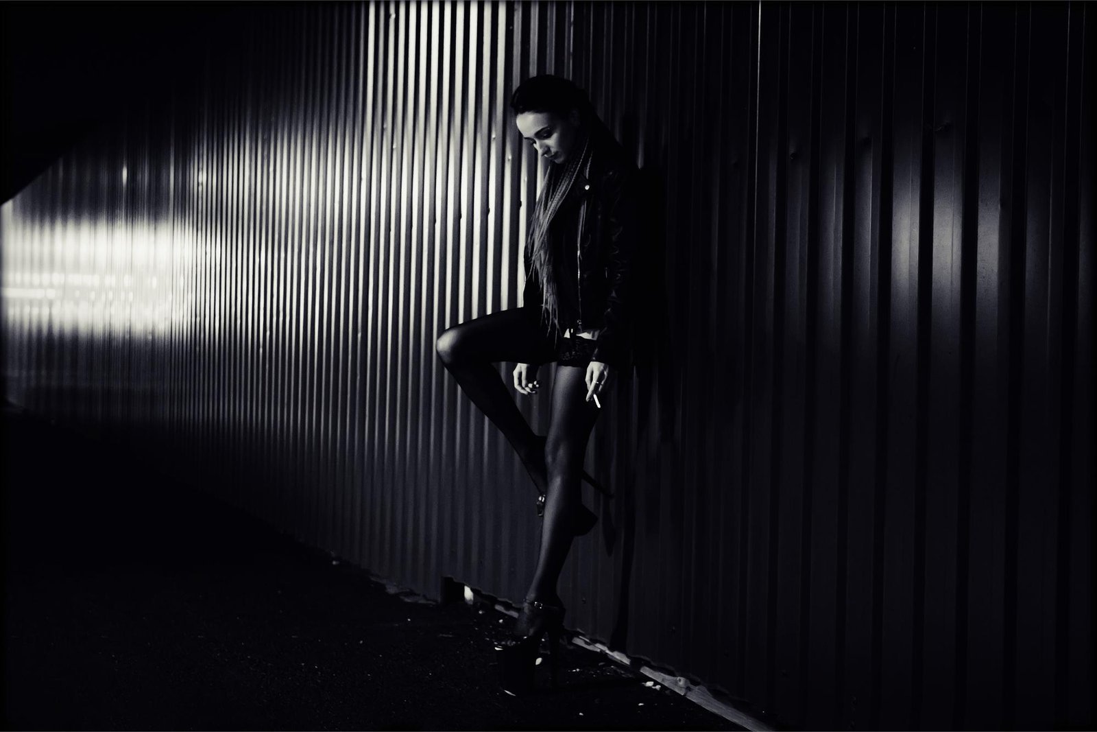 Woman in a leather jacket leaning against a metal wall in dim lighting, embodying a moody, edgy street style.