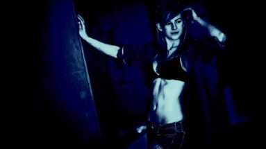 Woman in a dark urban setting, wearing a bra and open jacket, with jeans.