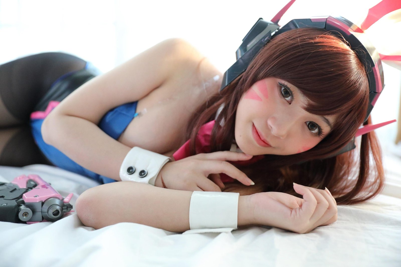 A close-up of a cosplayer lying on a bed, wearing a futuristic blue and pink costume with headphones, smiling softly while resting her hand on her chin, exuding a playful vibe.