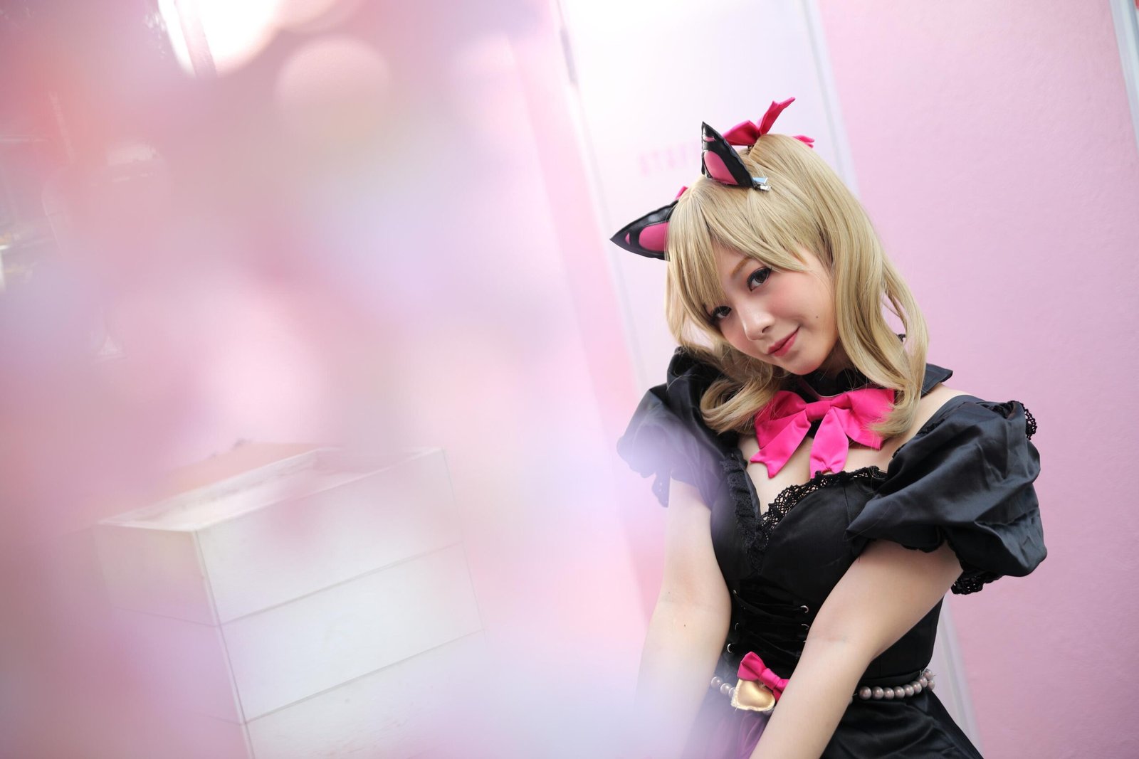 A cosplayer with blonde hair, dressed in a cute cat-themed outfit with black cat ears, a pink bow, and a black dress, posing in a soft, pastel-colored environment.