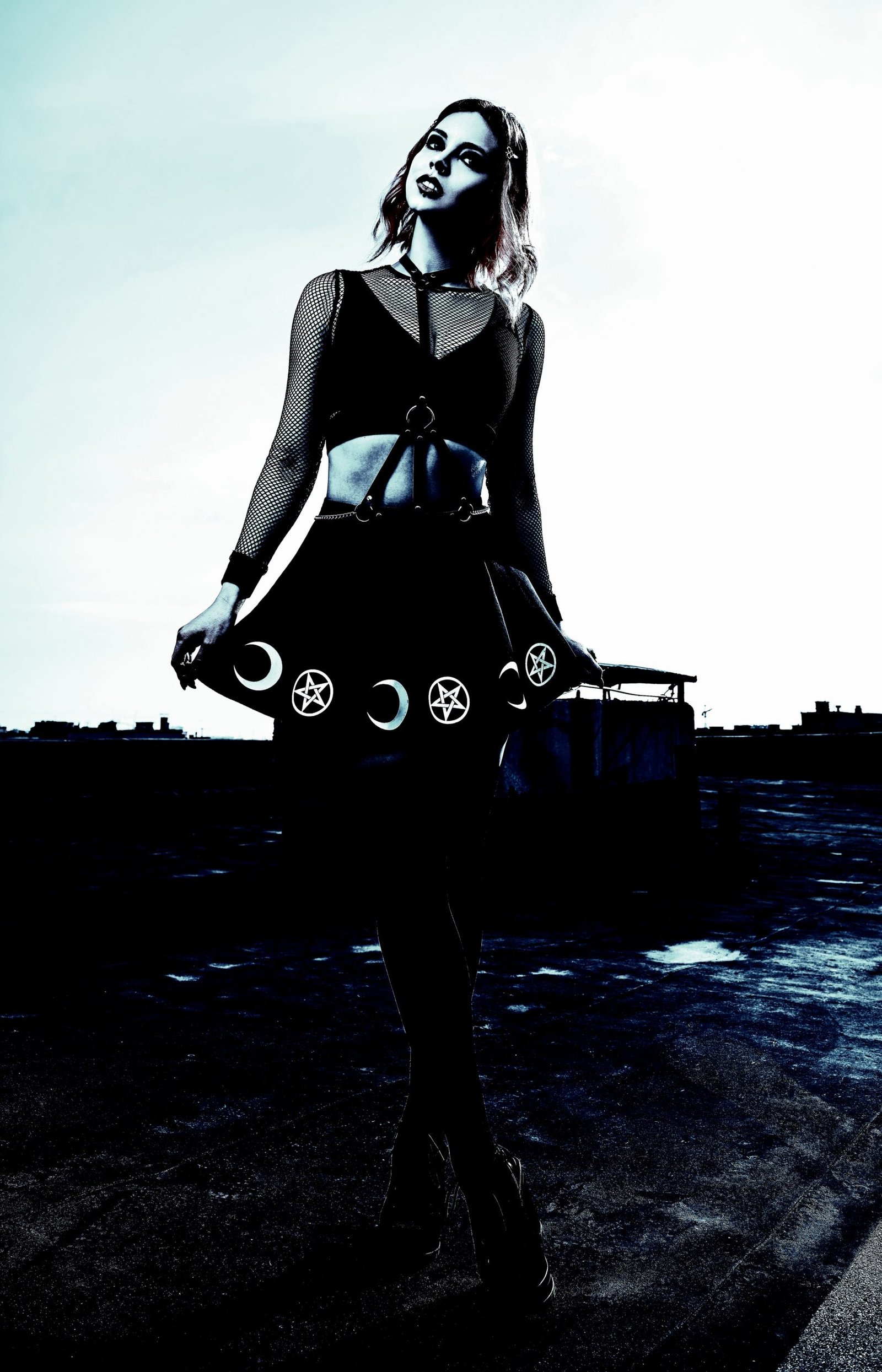 A woman in a gothic outfit standing on a rooftop, wearing a black skirt adorned with pentagram and moon symbols, against a cloudy sky.