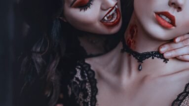 Vampire woman biting another woman's neck, showcasing dramatic makeup and gothic lace choker.