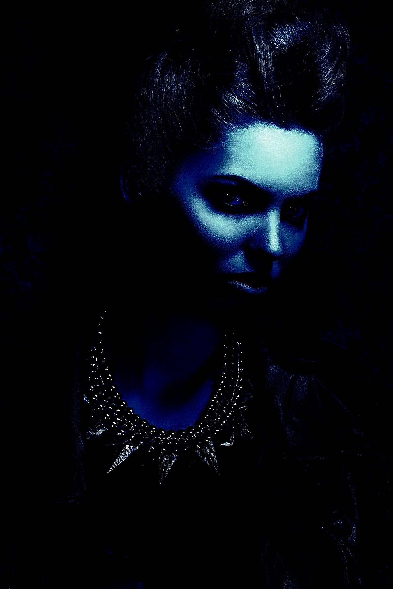 Woman with an intense gaze wearing a spiked gothic necklace under dim blue lighting, exuding dark elegance and mystery.