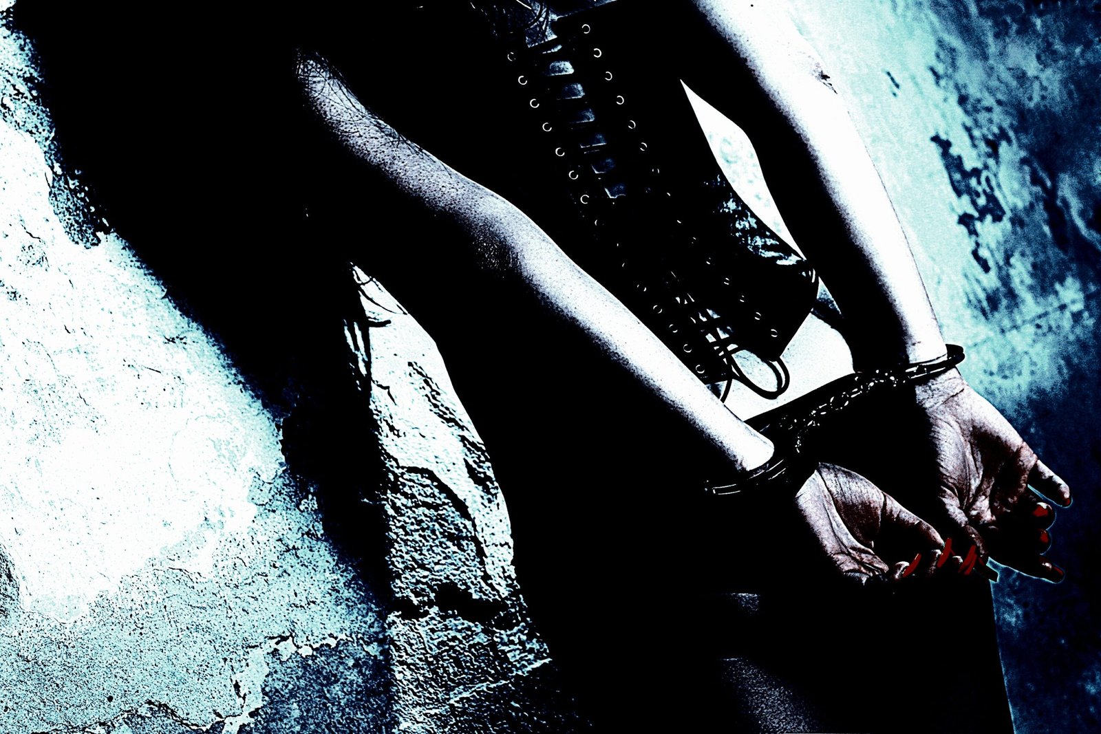 A person in a black corset with hands handcuffed behind their back, leaning against a wall.