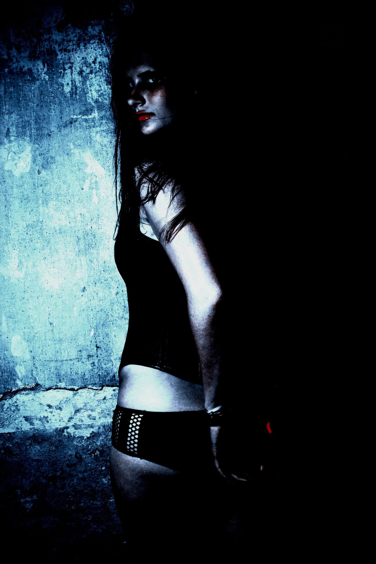 Woman in black lingerie, standing against a grungy wall, partially illuminated in a dark room.