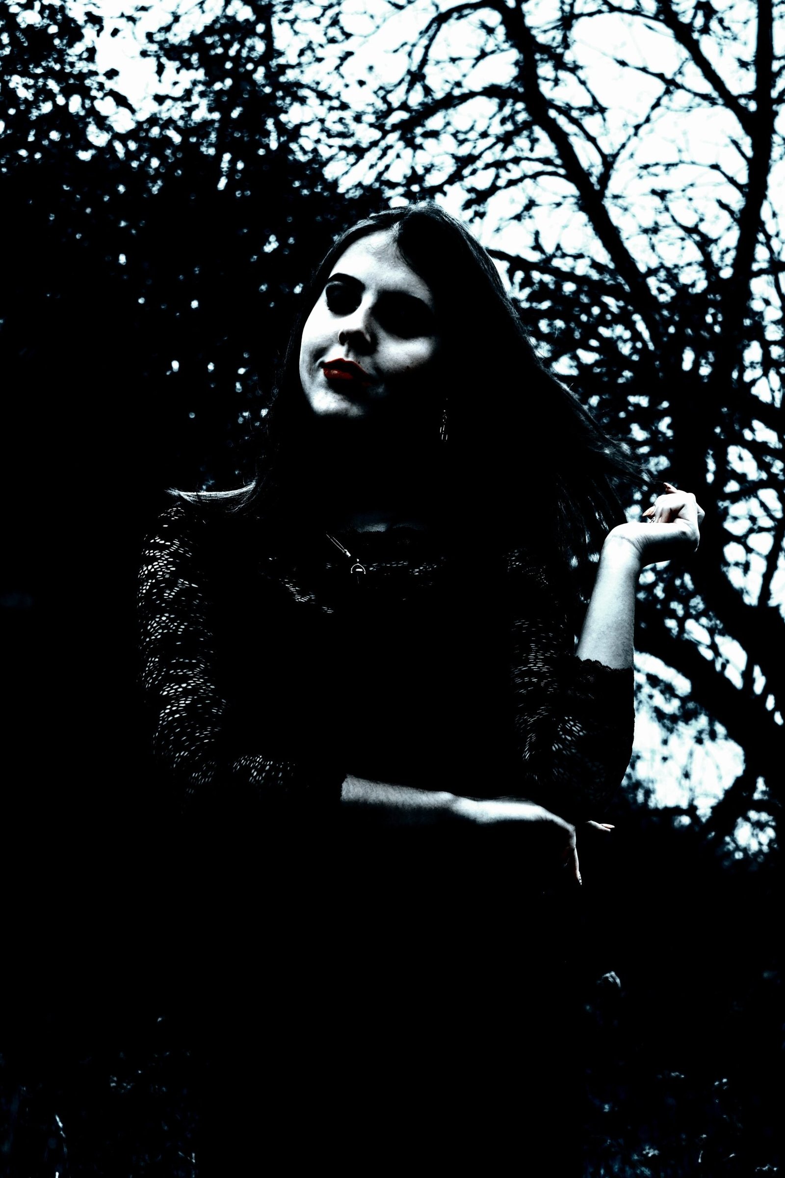 A gothic woman standing outdoors with dark makeup and red lips, wearing a black lace outfit against a backdrop of twisted, leafless trees.