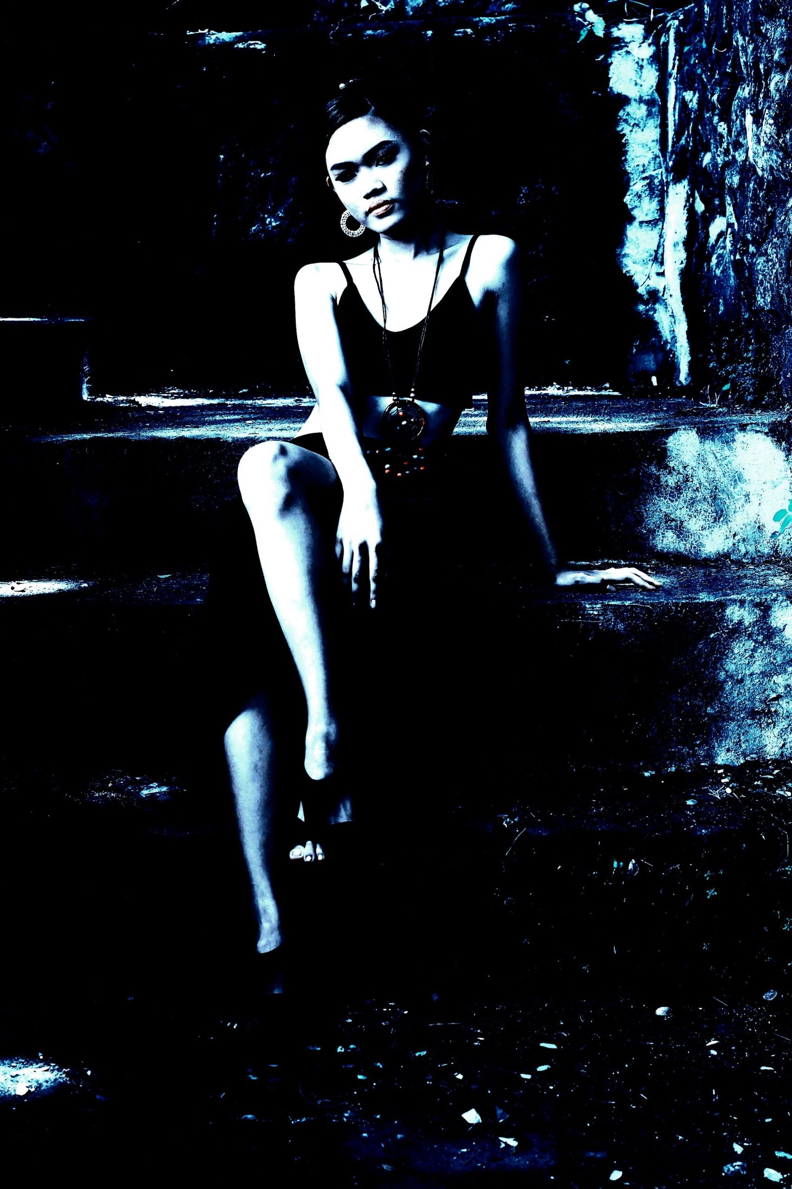 A woman in gothic attire sitting on steps, wearing a black outfit with a large pentagram necklace, set against a dark, textured background.