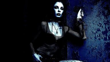 Gothic woman in dark clothing, adorned with chains and piercings, standing in a mysterious, shadowy environment.
