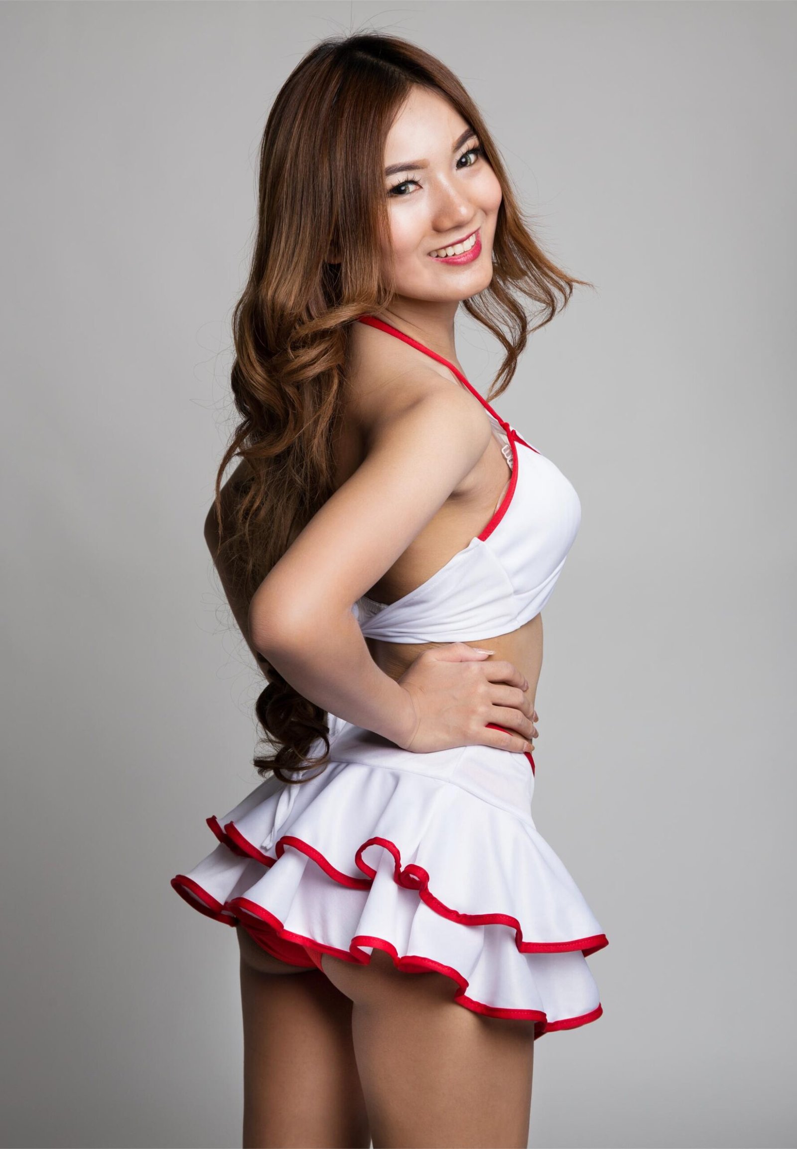 A smiling woman in a white and red cheerleader-inspired costume, posing against a light gray background, showcasing a playful and confident expression.