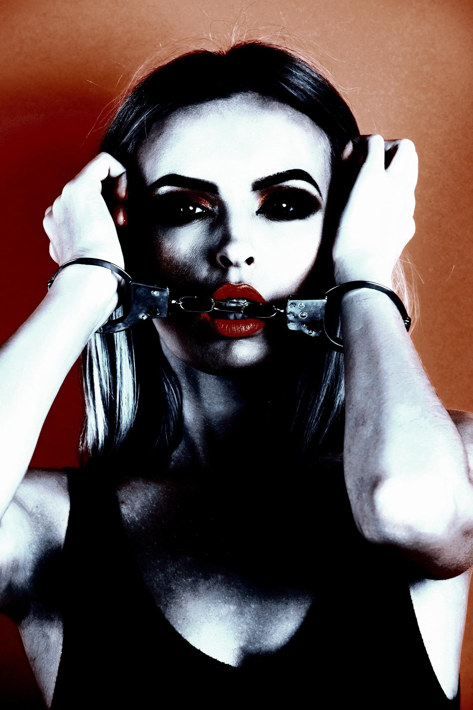 Woman with bold red lips, holding handcuffs to her face against a striking orange backdrop, creating a powerful, intense look.