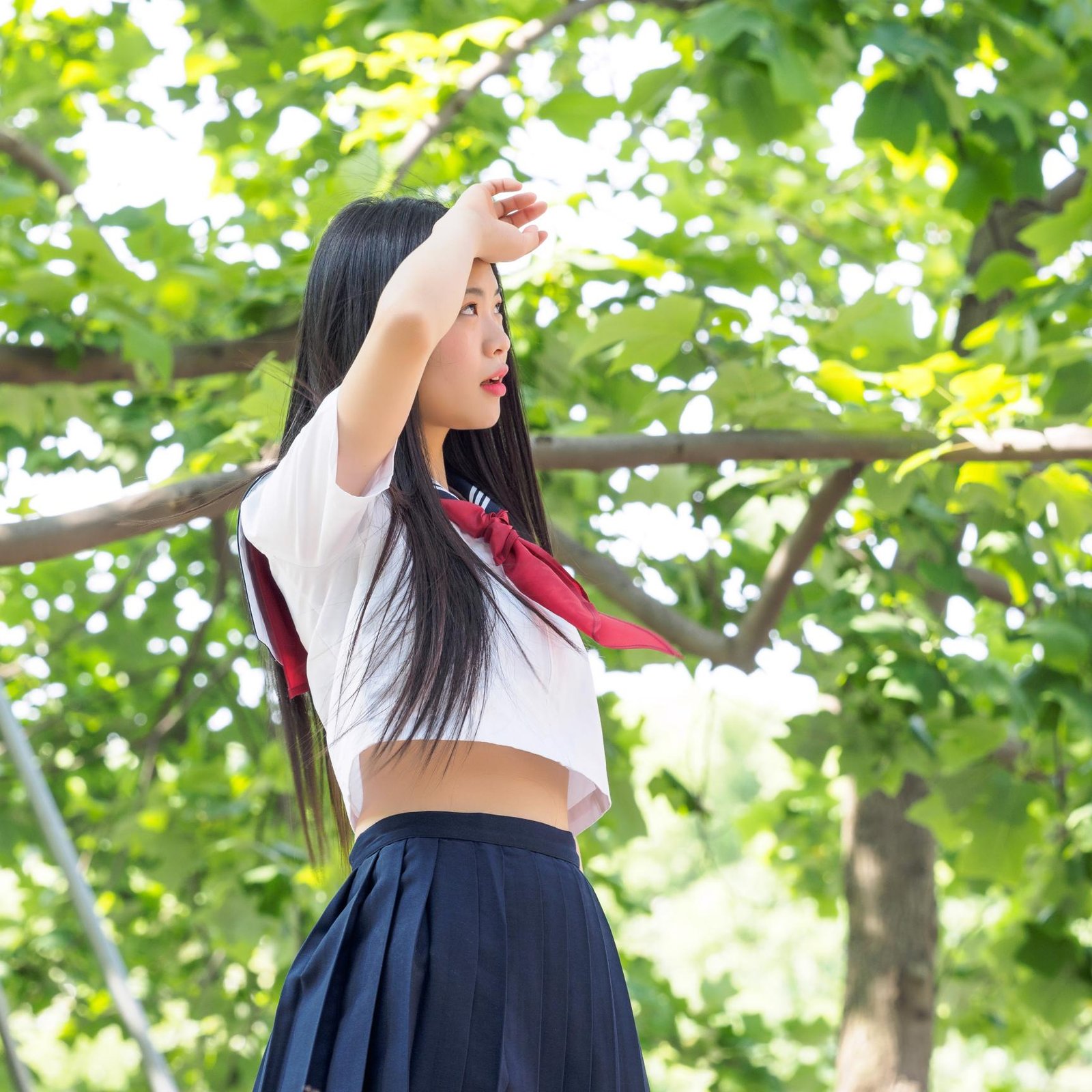 A young woman in a school uniform with long black hair, looking into the distance while standing outdoors among lush green trees.