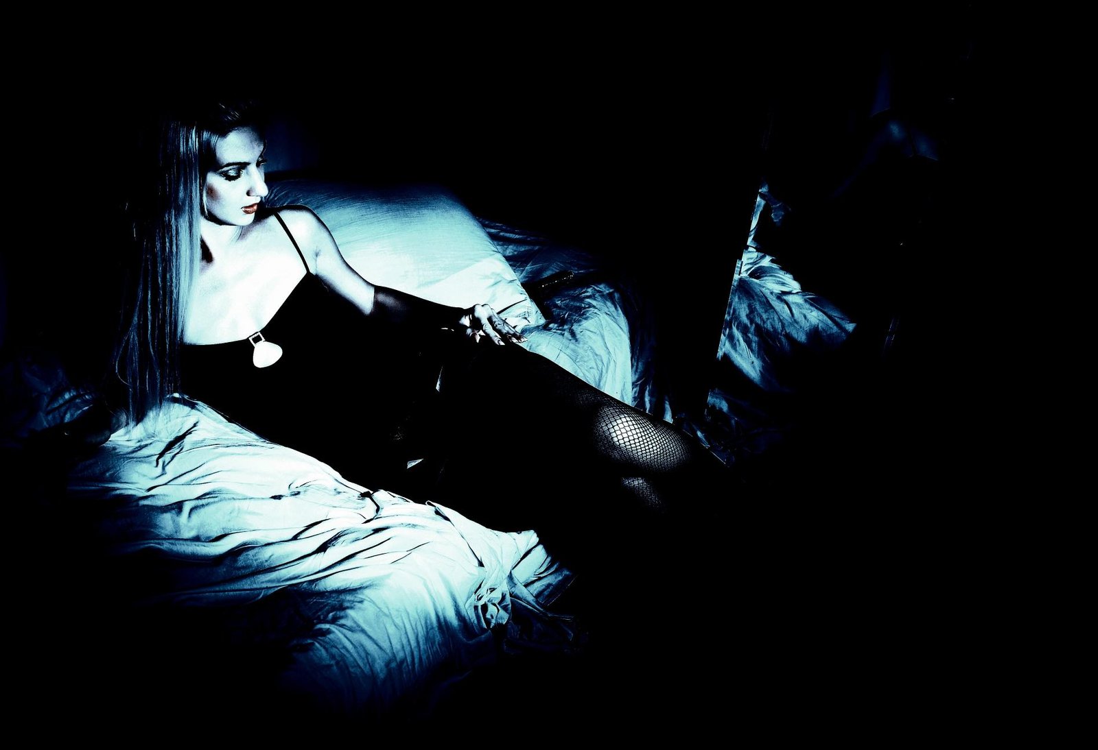 Woman in black dress reclining on a bed in low lighting, capturing a sultry, dark atmosphere.
