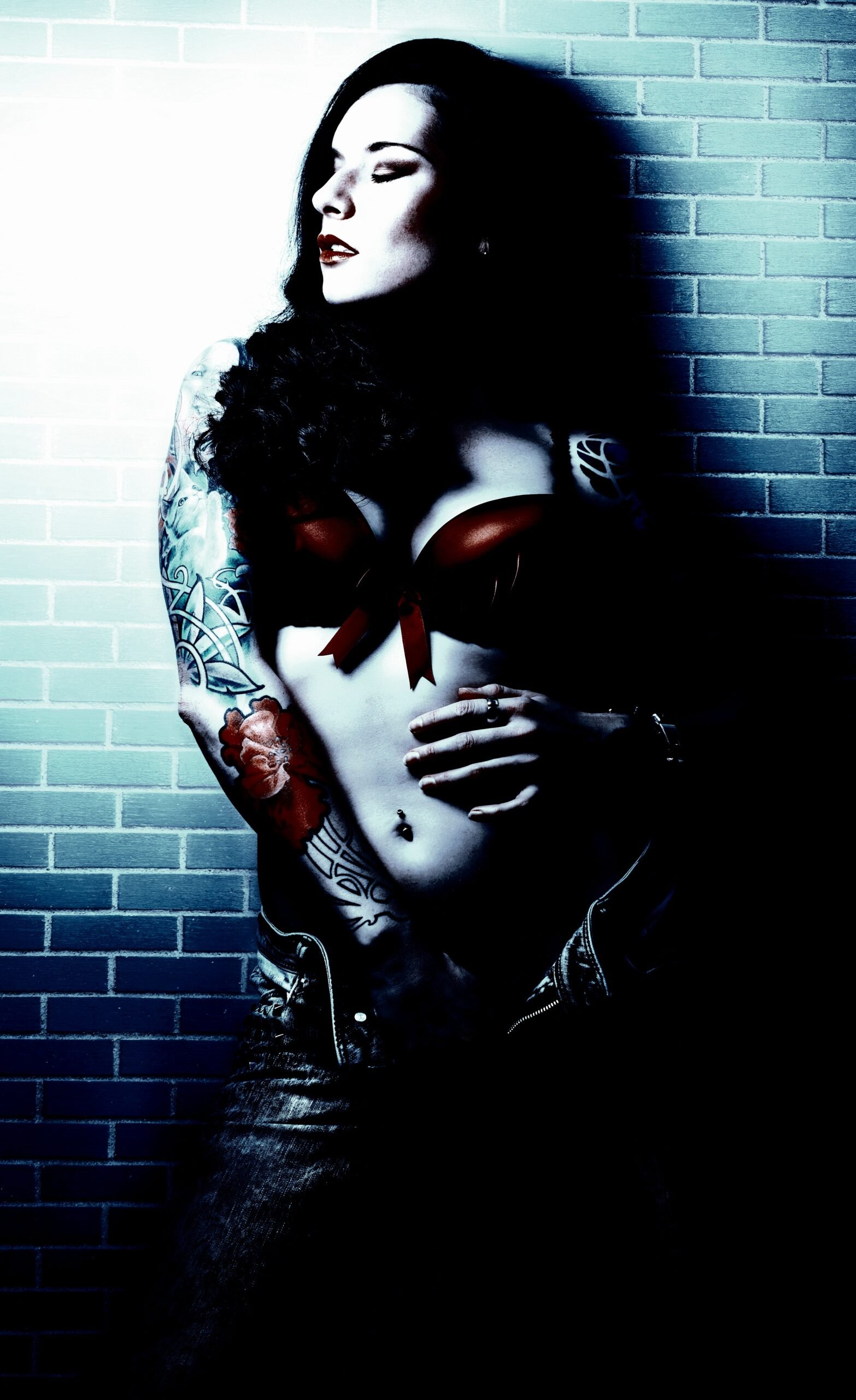Gothic woman with intricate tattoos and a red bra, posing against a brick wall.