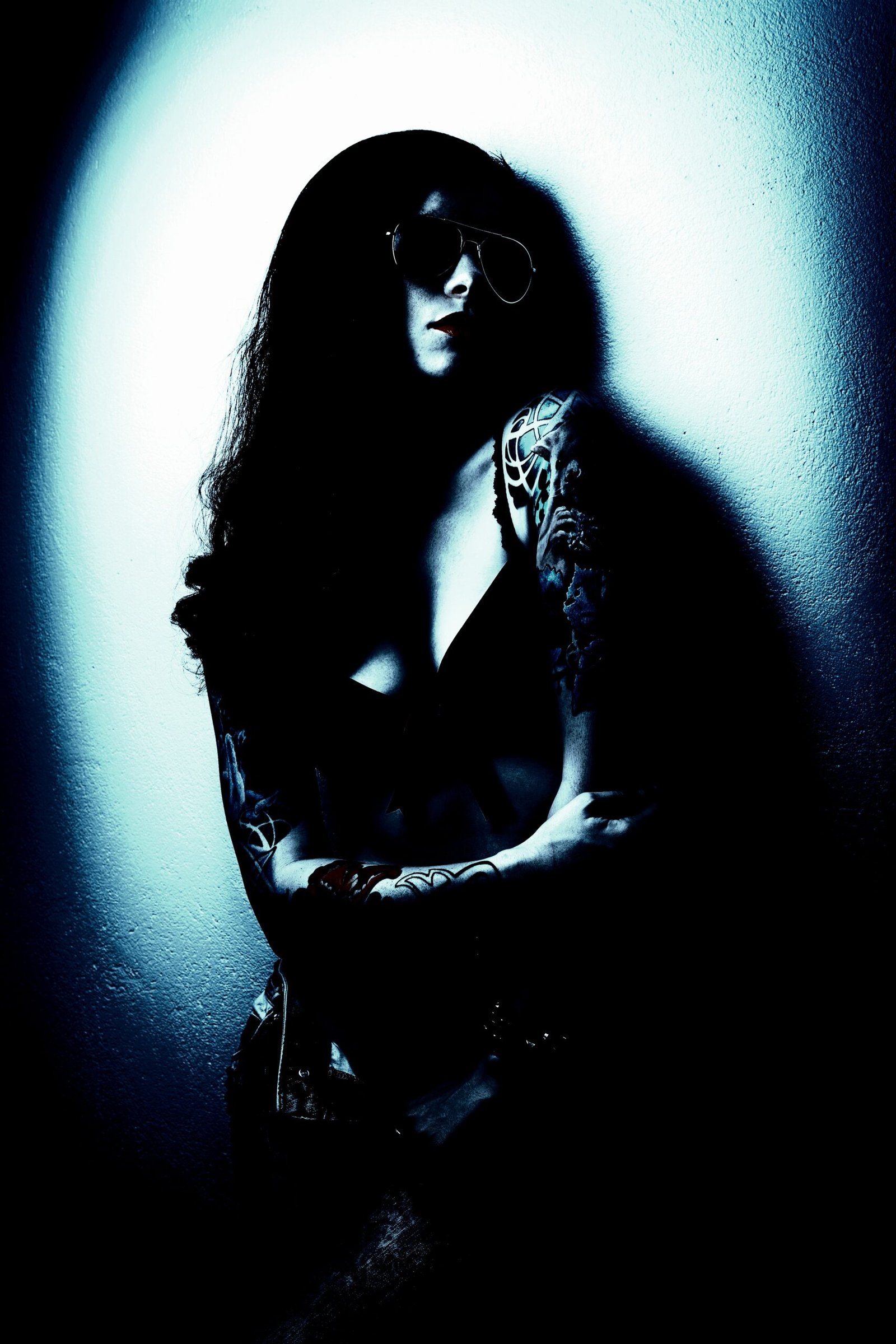 Mysterious gothic woman with tattoos and sunglasses, standing against a light-colored wall.