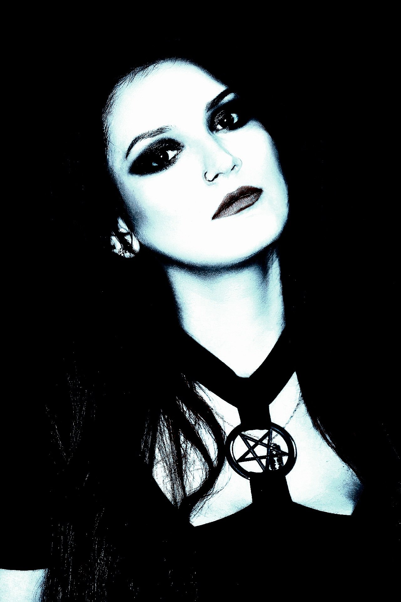 A young woman with dark makeup and a gothic outfit, standing outdoors with a serious expression, holding her hair.