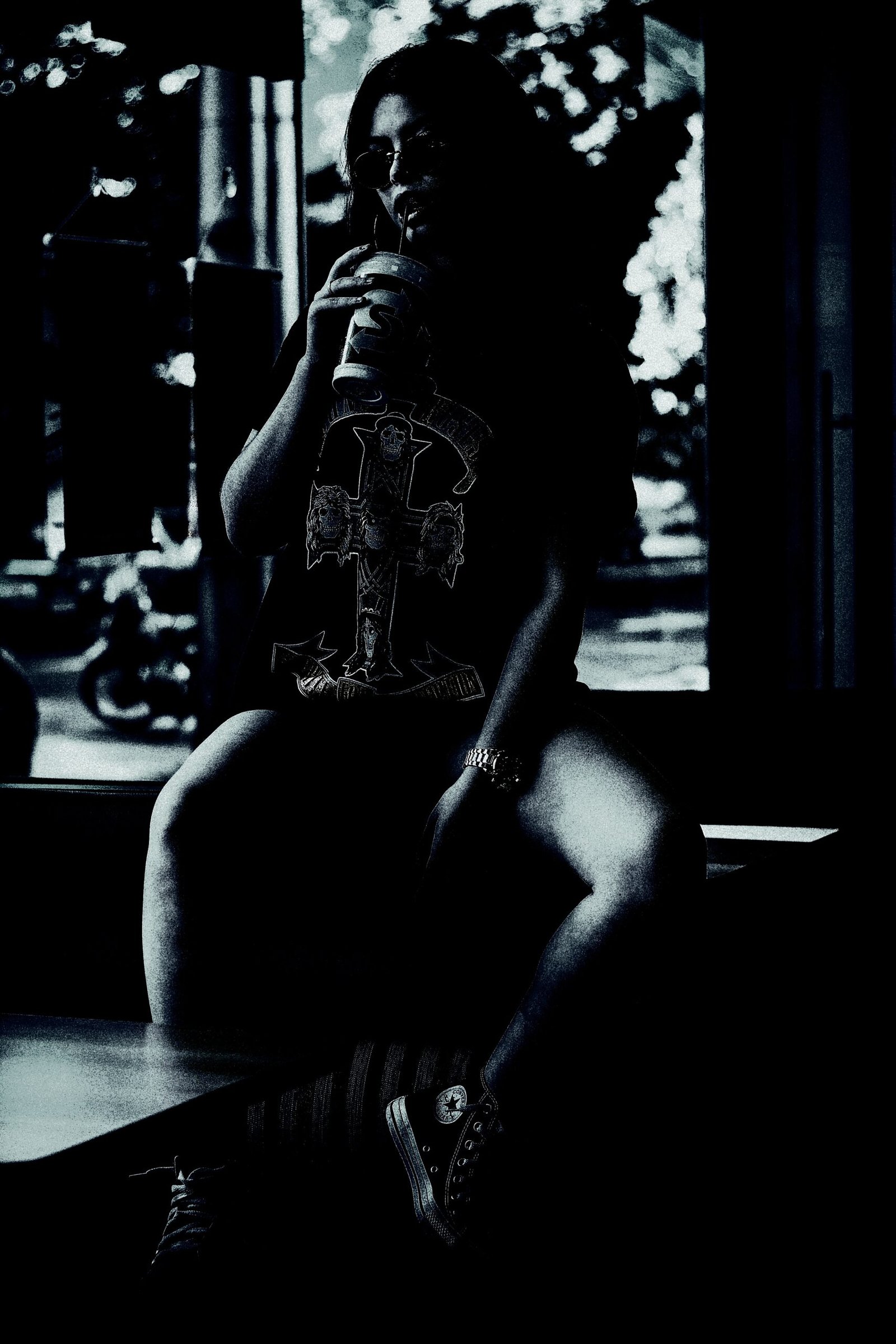 Gothic woman sitting in a dimly lit room, sipping a drink, wearing a graphic tee with a dark, occult-inspired cross design.