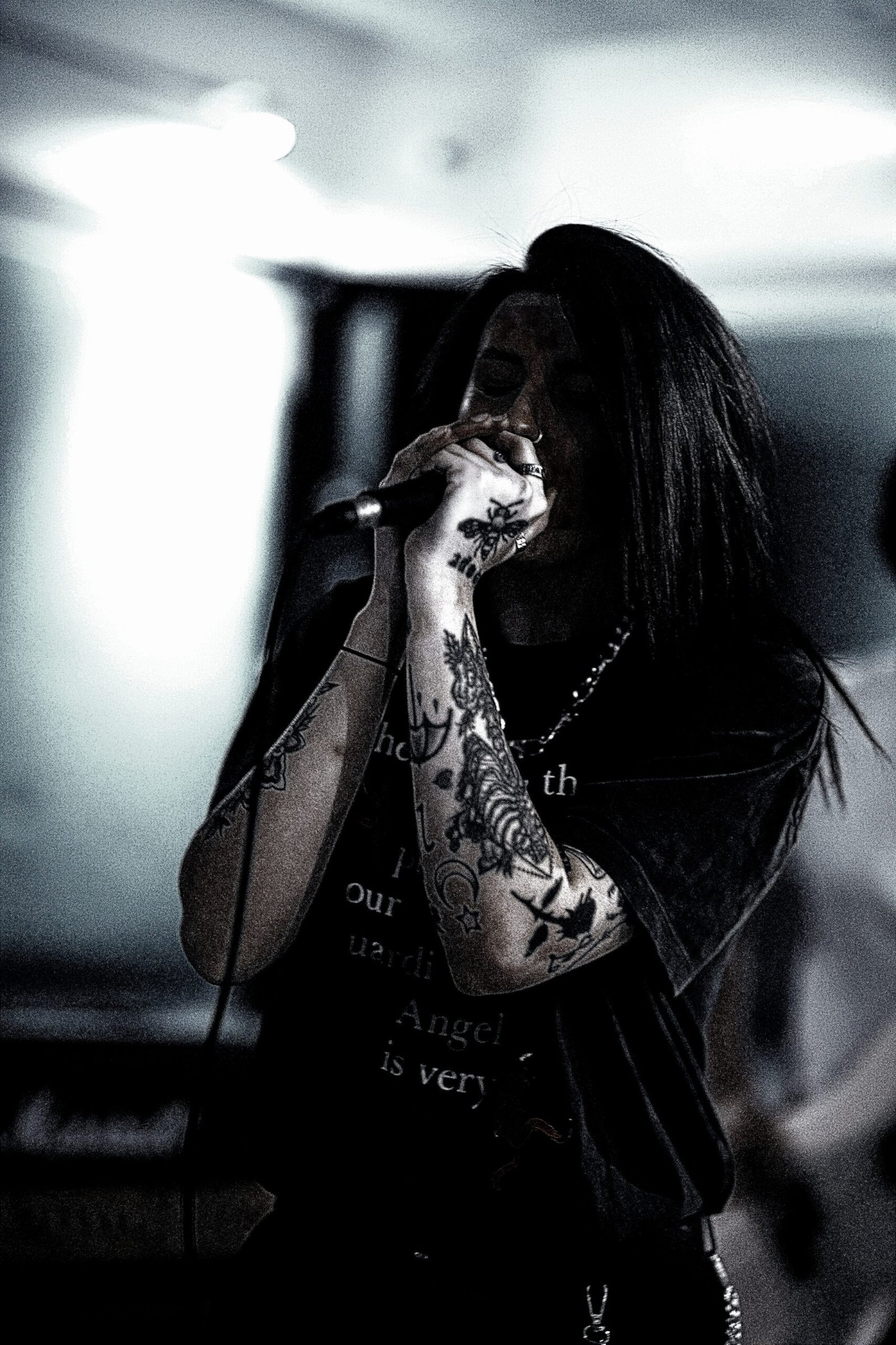Close-up of a tattooed female singer gripping a microphone passionately, performing in a low-lit, intimate concert setting.