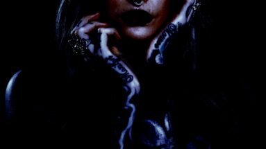 Close-up of a gothic woman with dark lipstick, septum piercing, and tattooed hands in a moody, shadowy setting.