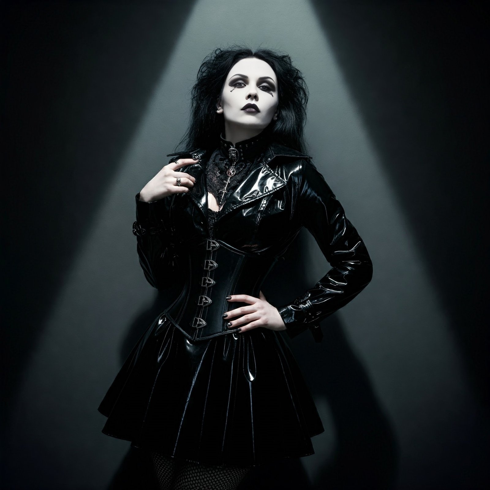 A gothic woman in a black PVC outfit, standing under dramatic lighting in a studio, with her hands on her hips and a serious expression.