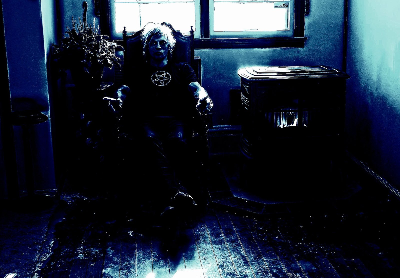 A gothic man sitting in a dimly lit room, wearing a pentagram shirt, with a fireplace casting eerie shadows, creating a dark and mysterious atmosphere.