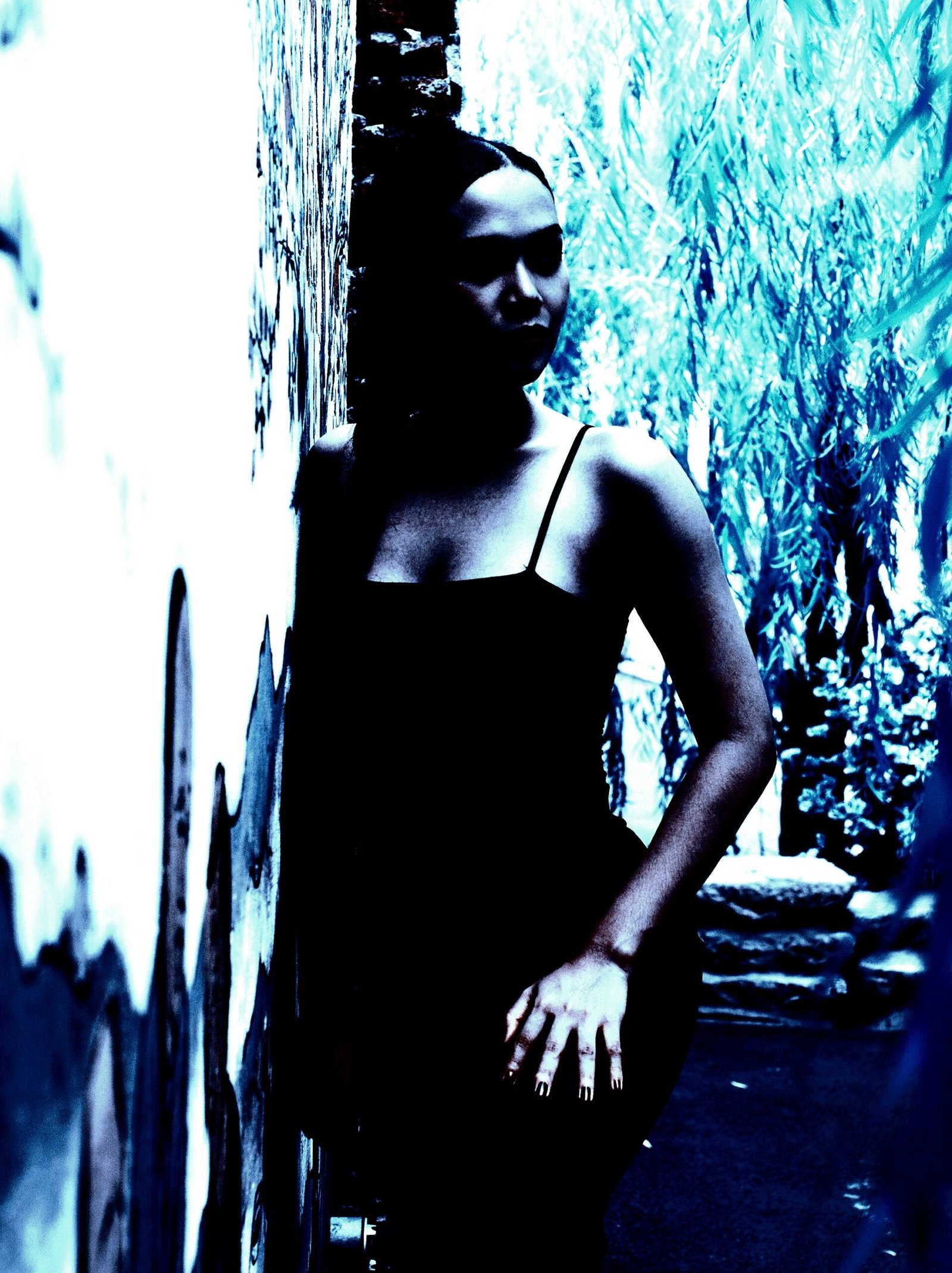 Woman in a black dress standing against a graffiti-covered wall, surrounded by moody blue-green foliage.