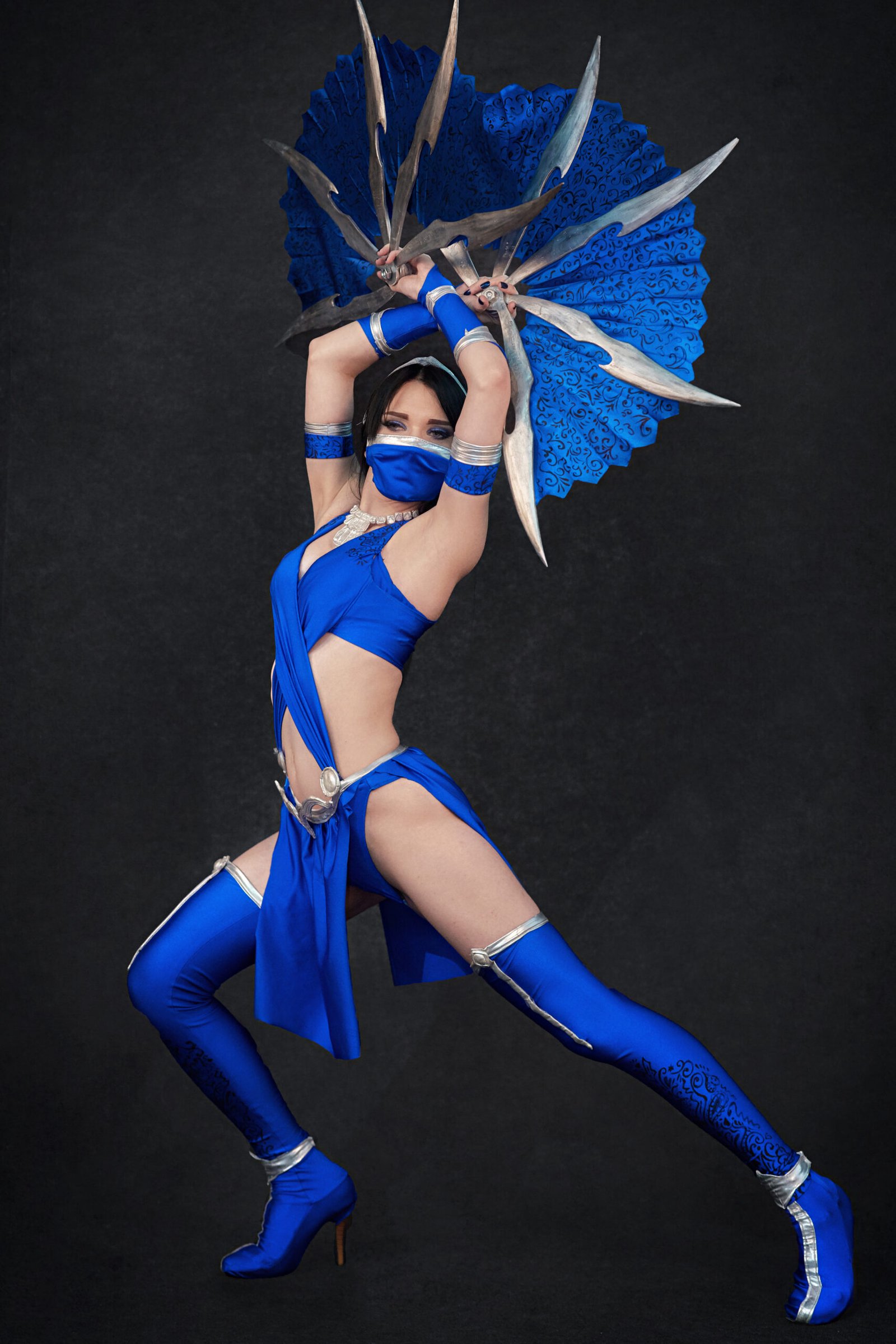Cosplayer dressed as Kitana from Mortal Kombat, posed dynamically while holding steel fan blades.