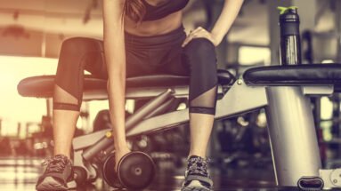 Woman in a gym lifting a dumbbell while sitting on a bench, bathed in warm sunlight.