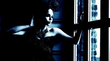 Woman in a gothic lace top and ornate necklace, gazing out a window with a contemplative expression in soft blue lighting.