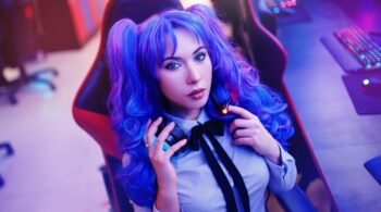Woman with blue-purple hair in pigtails, seated in a gaming chair, holding headphones with a focused expression.