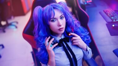 Woman with blue-purple hair in pigtails, seated in a gaming chair, holding headphones with a focused expression.