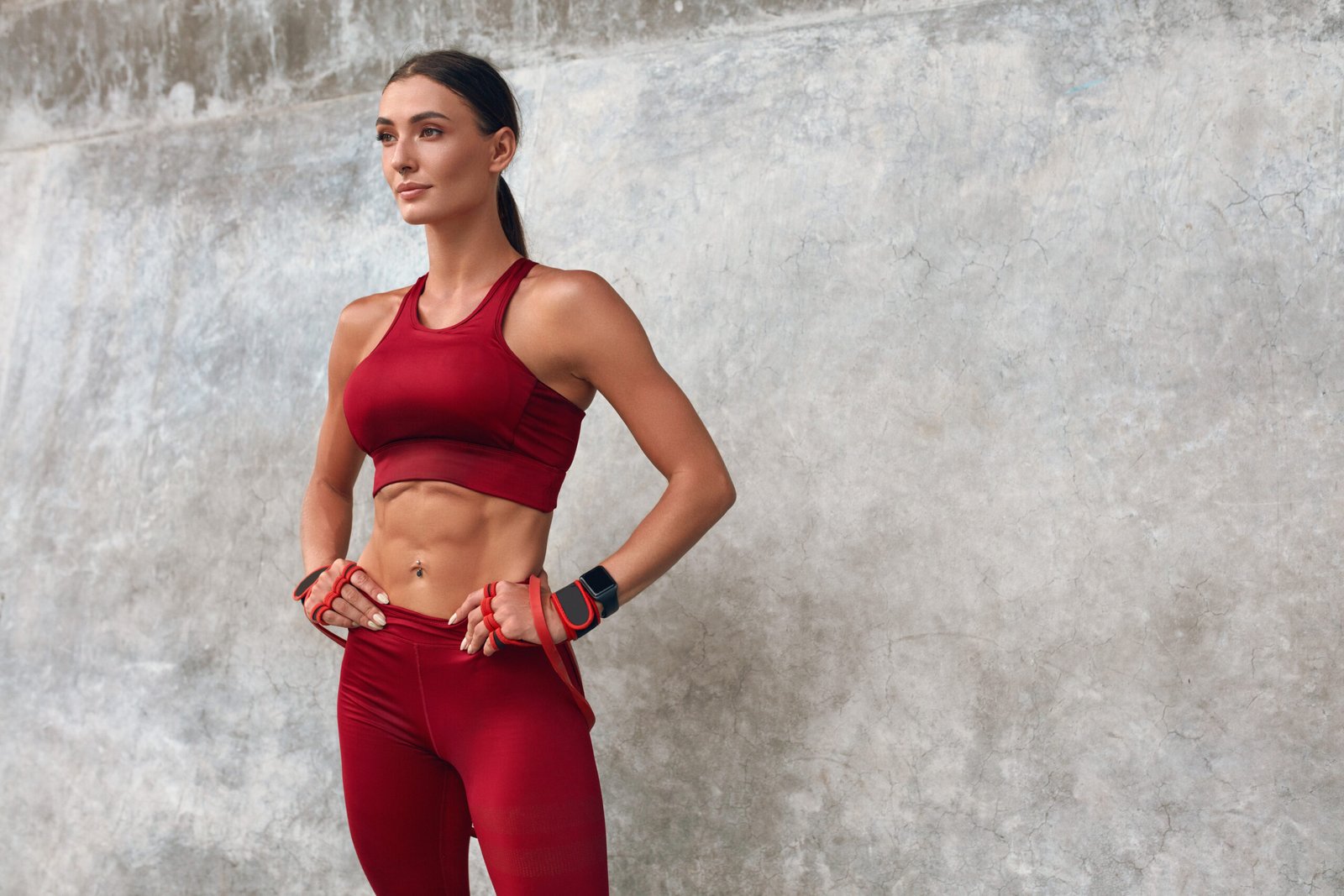 Fit woman in a red sports outfit with hands on her hips, standing against a textured concrete wall.