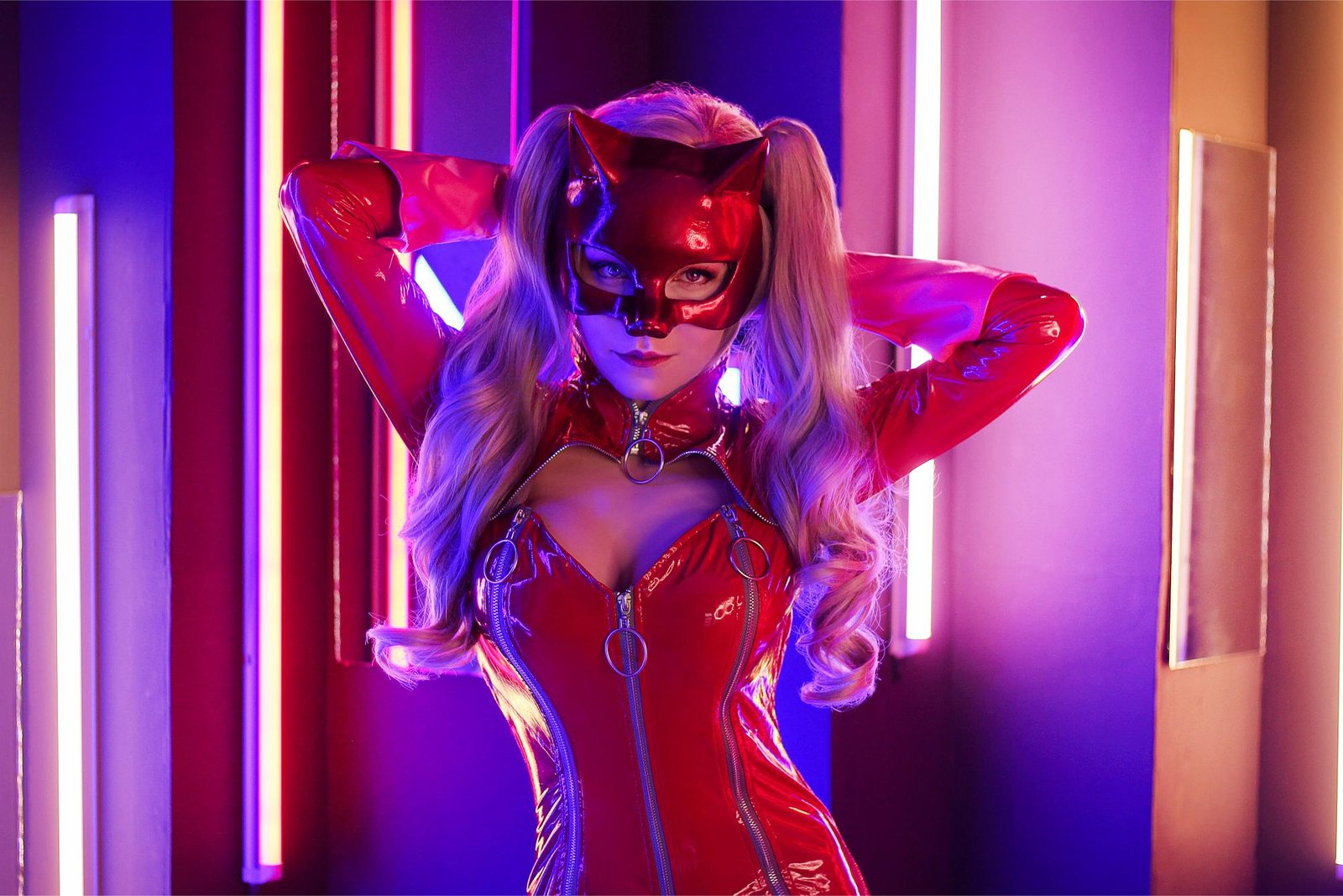 Cosplayer in red latex cat suit and mask, posing with hands behind head in a vibrant neon-lit room.