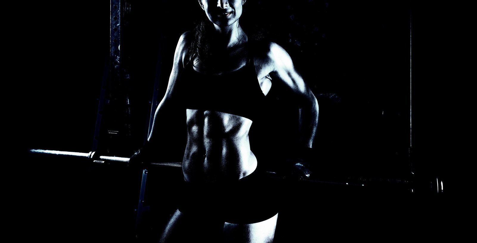 Fit woman showcasing muscular definition while gripping a barbell in a dark gym environment.
