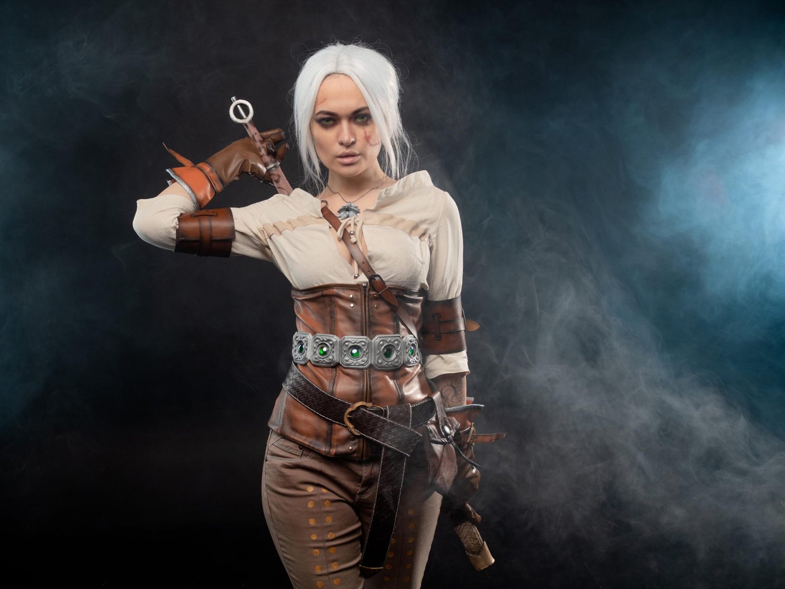 Cosplayer dressed as Ciri from The Witcher with white hair, leather armor, and a sword handle, standing against smoky background.