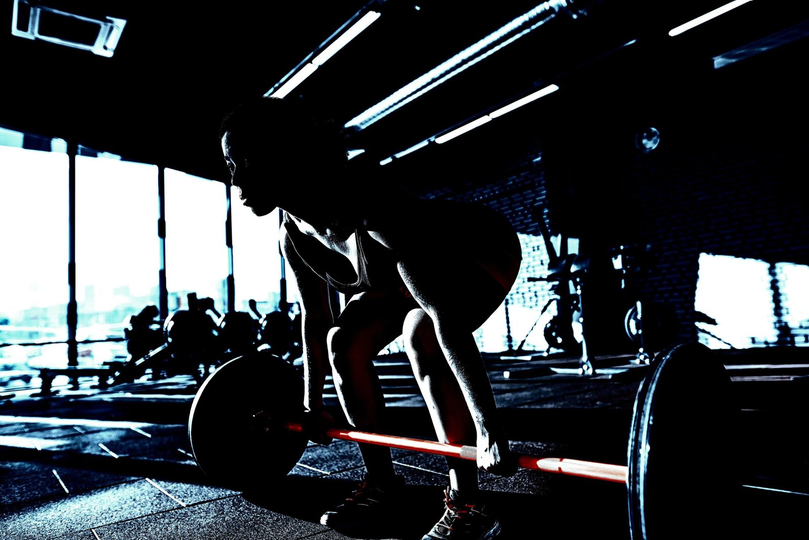 Silhouette of a woman performing a deadlift in a gym, framed by natural light streaming through windows.
