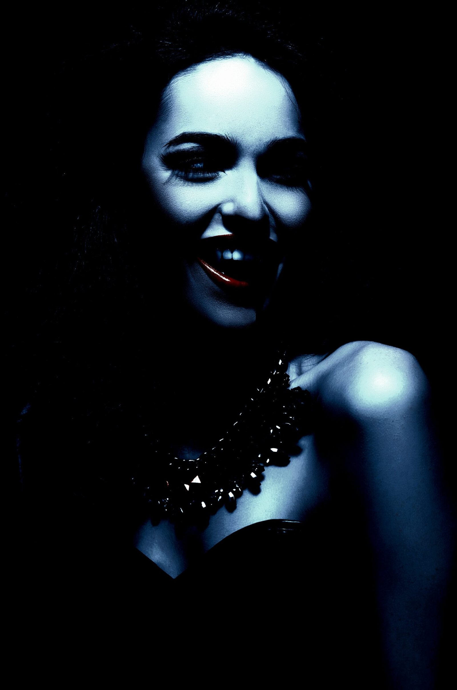  Woman smiling enigmatically, her face partly shadowed, wearing a dark, intricate necklace on bare shoulders.