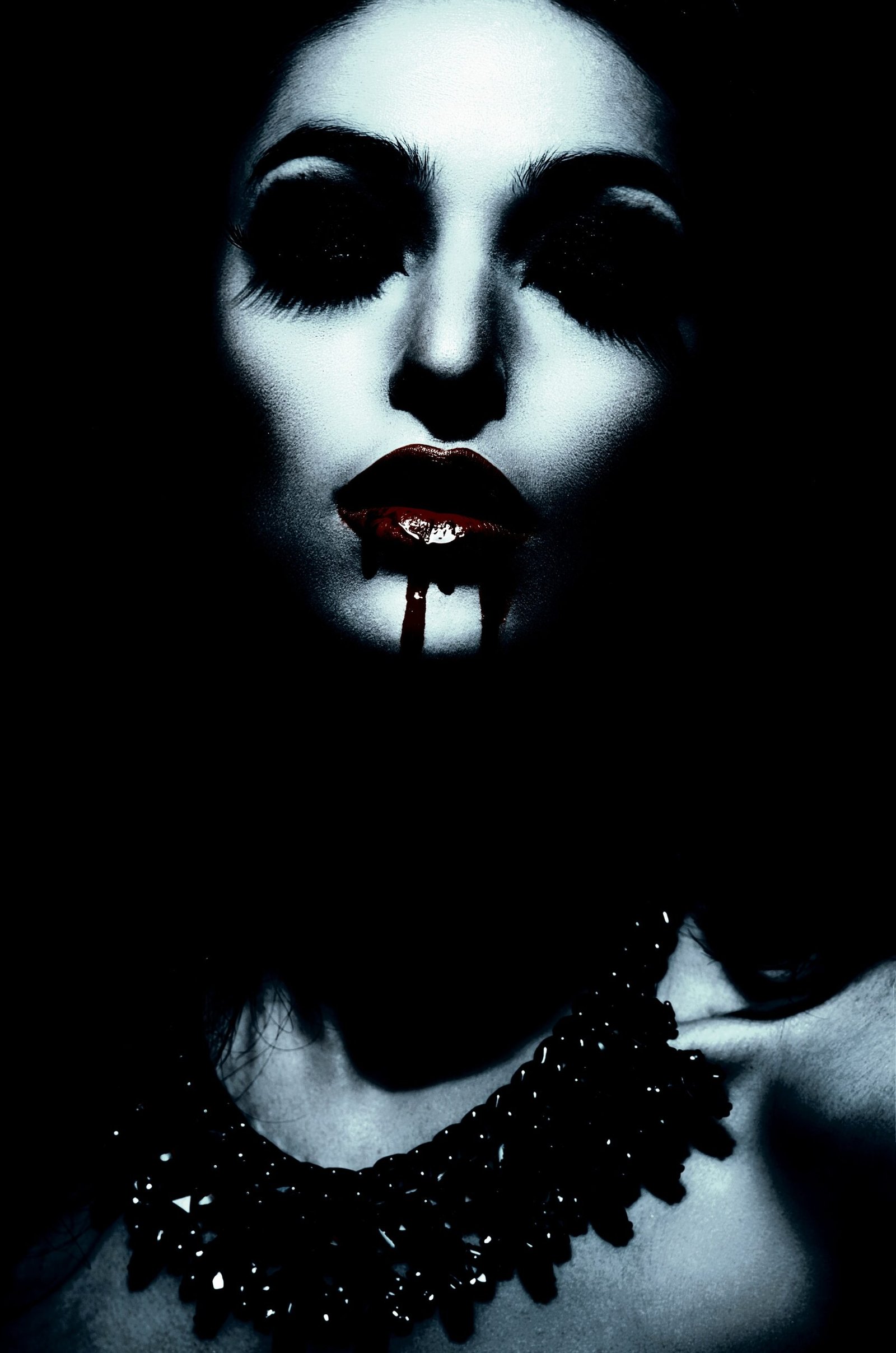 Close-up of a woman with dark makeup and blood-red lipstick, wearing a bold, gothic necklace against her pale skin.