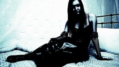 Woman sitting on a bed wearing a gothic black dress, with fishnet gloves and a mysterious expression.