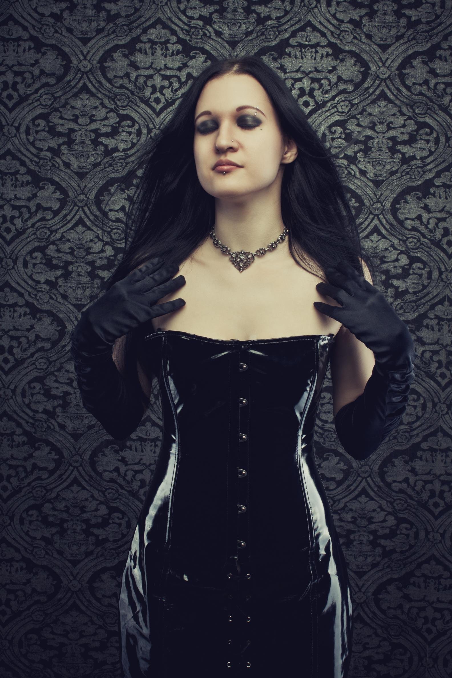Gothic-inspired woman in a black latex corset and long gloves, posed against a textured dark background.