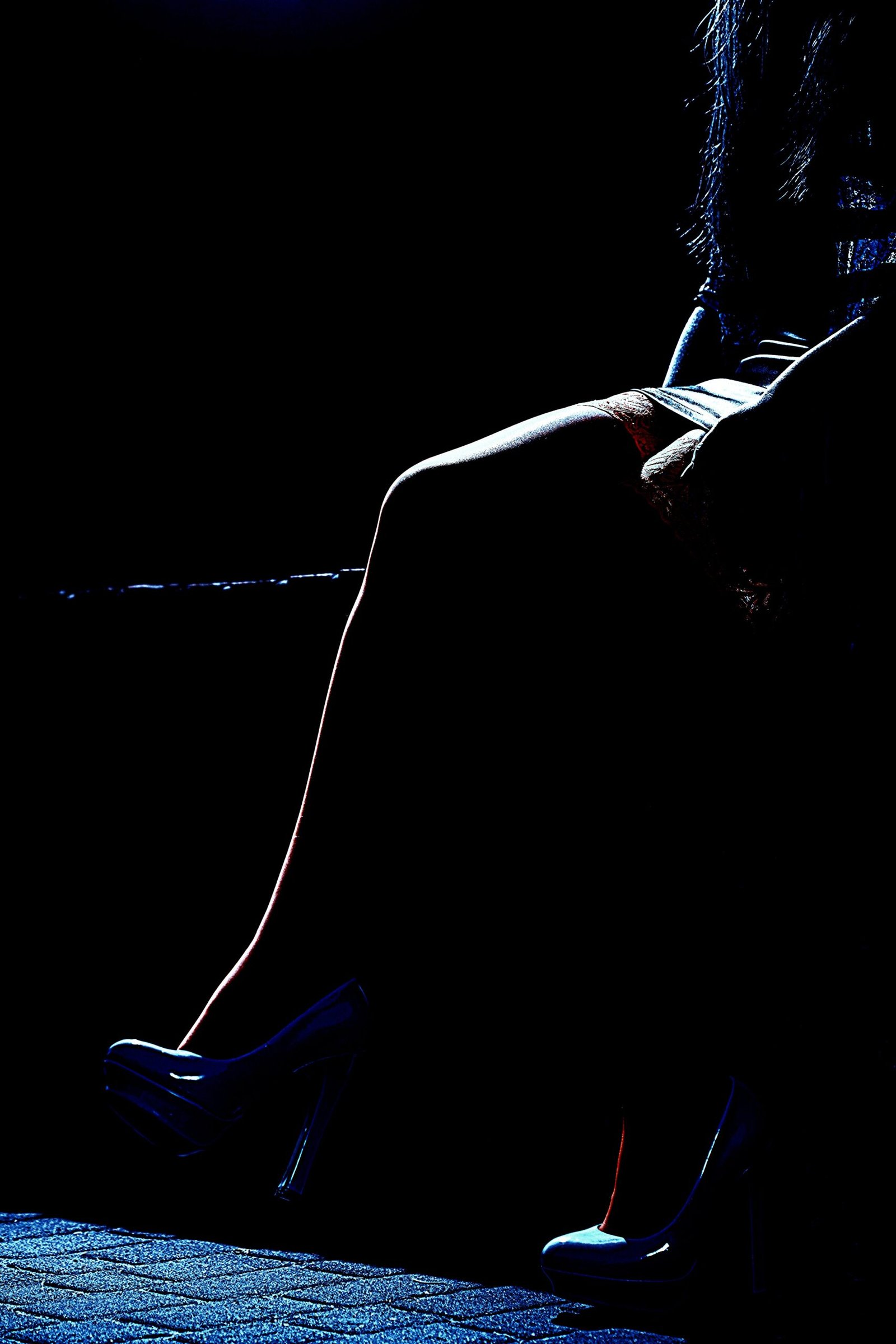 The legs of a woman in high heels, sitting in a dark, moody environment, with the focus on the curve of her leg and the reflective surface of her shoes.