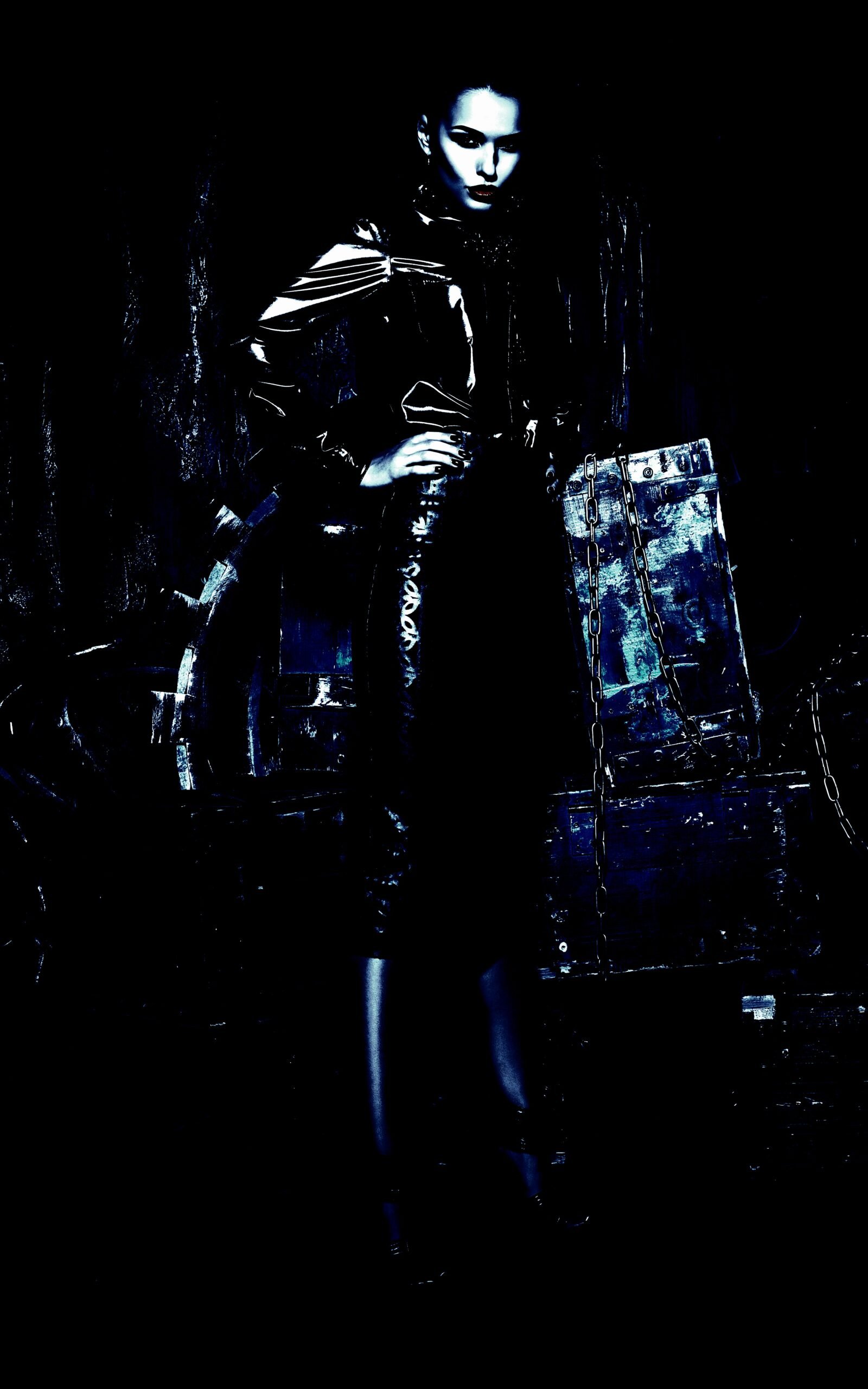 Gothic woman standing confidently in dark attire, surrounded by chains and industrial elements in a dimly lit scene.