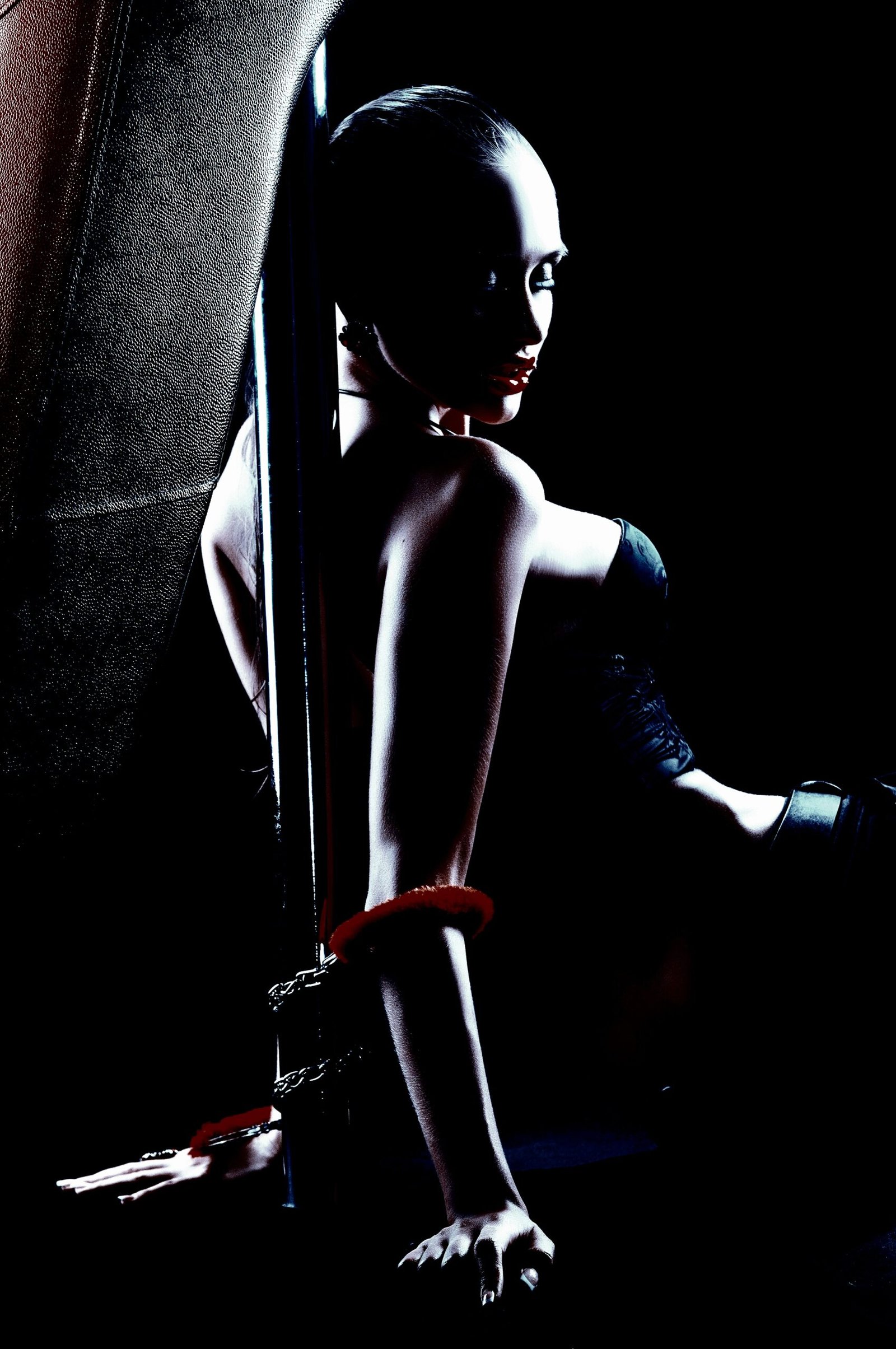 A woman in a black corset and wrist cuffs, seated next to a metal pole in a shadowy, dimly lit environment, exuding an aura of mystery and dominance.