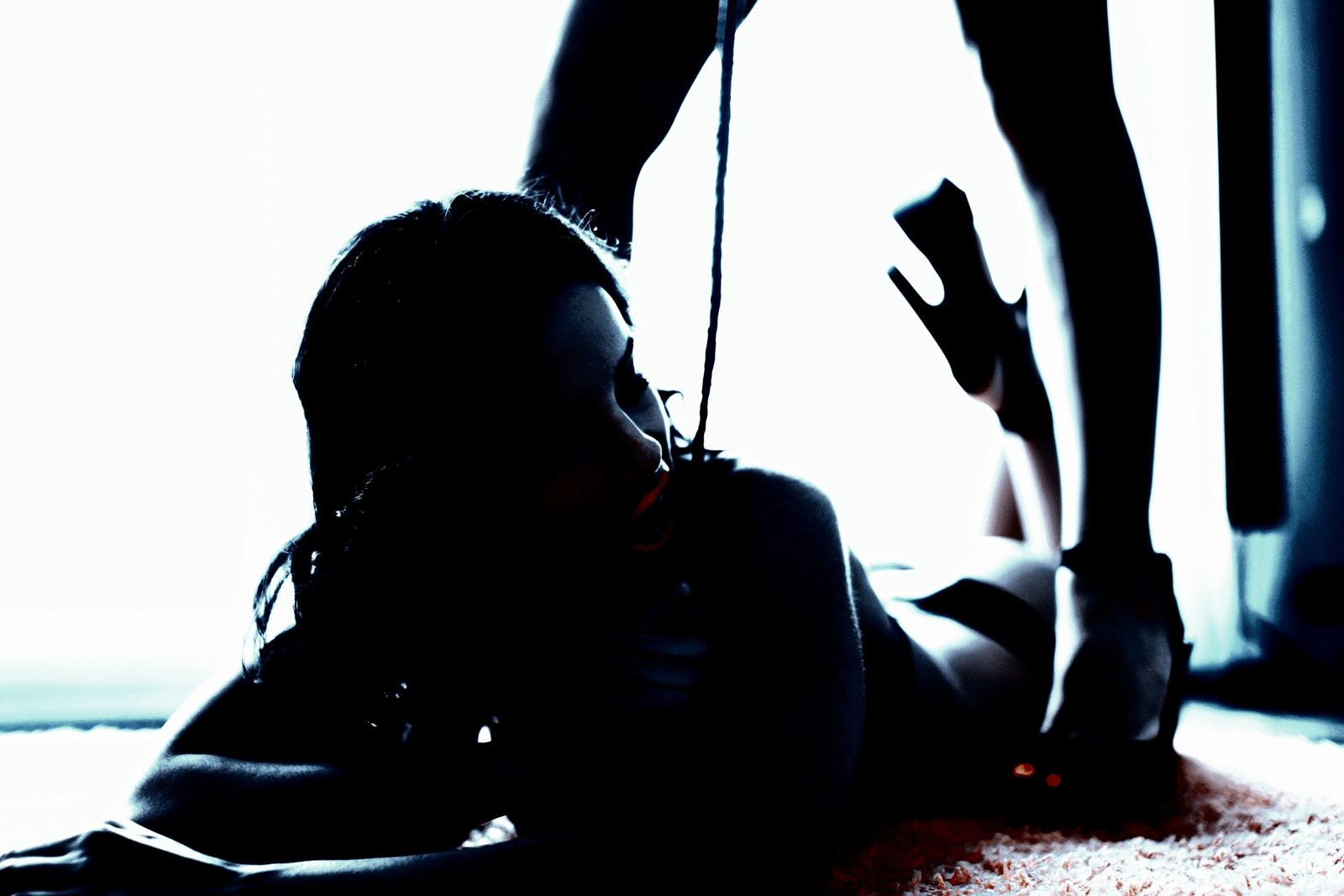 A dramatic, silhouette shot of a woman on the floor, submissive, while another person stands over her, their high heels highlighted in sharp contrast, creating a provocative and moody scene.