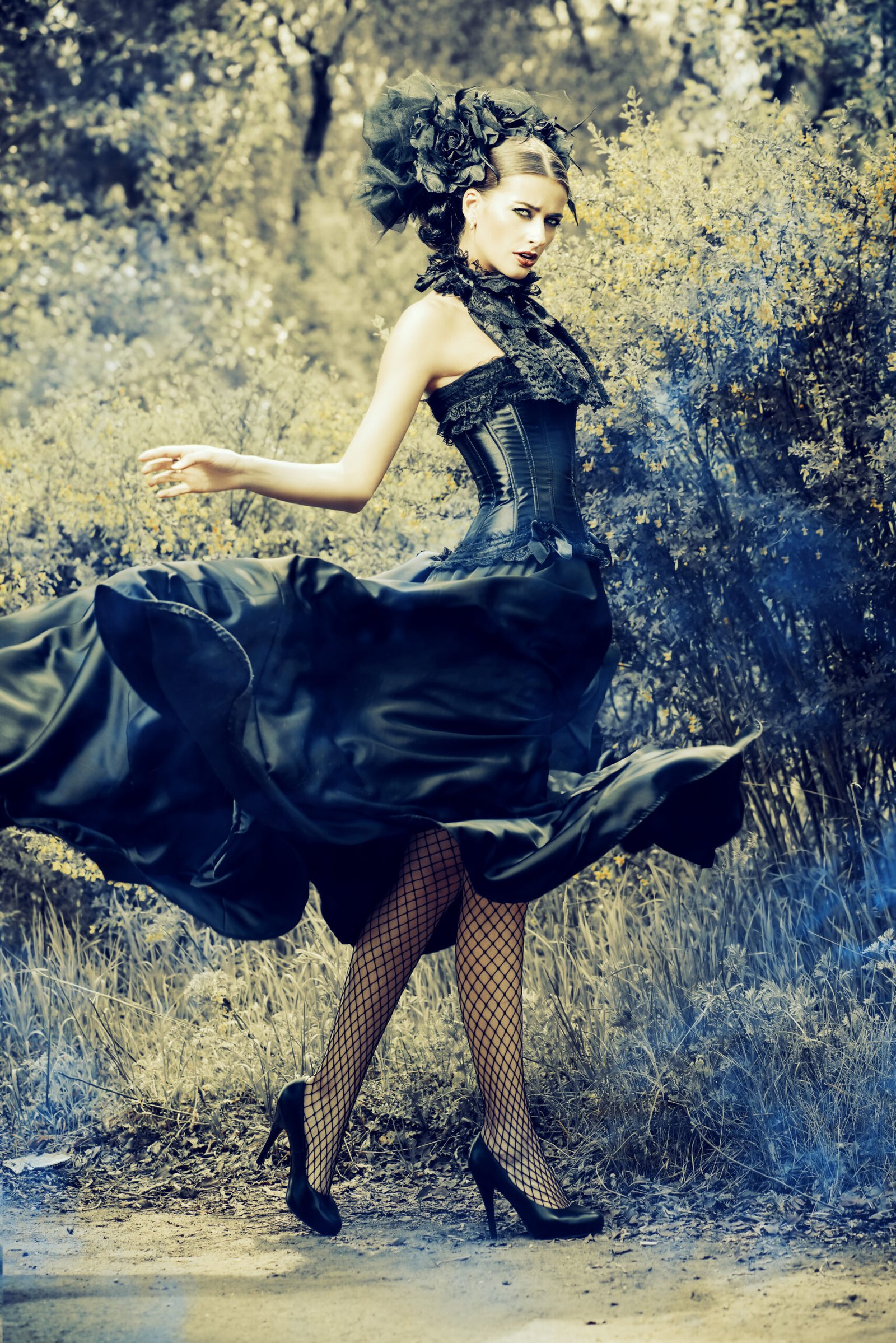 Woman in a flowing black goth cocktail dress with corset details, spinning dramatically in an outdoor setting, wearing fishnet tights and high heels.